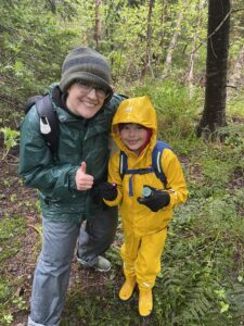 an adult and a child in raingear in the woods, giving a thumbs up